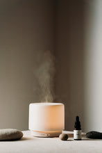 Load image into Gallery viewer, Cedar Aroma Diffuser with the lamp function on and the light shining through the ceramic cover. Sulis essential oil blend at the side with some pebbles for decoration